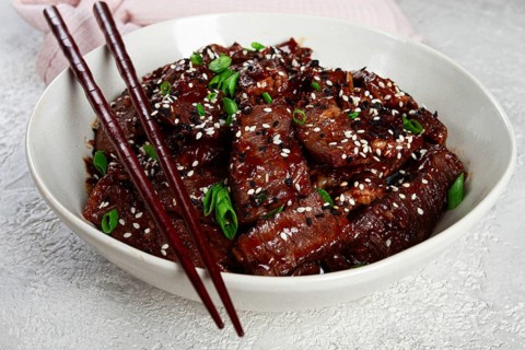 Crispy beef, with green onions and sesame seeds, in a bowl with chopsticks, on a textured surface,