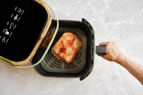 A hand removes an air fryer basket with roasted piece of pork fillet, top view