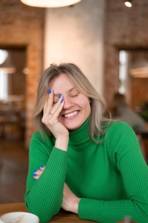A woman in a green sweater smiles warmly while enjoying breakfast at a cozy cafe. The ambient