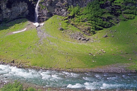 A clear river flows through a green landscape with a waterfall and rocky hills, Rallarveien,