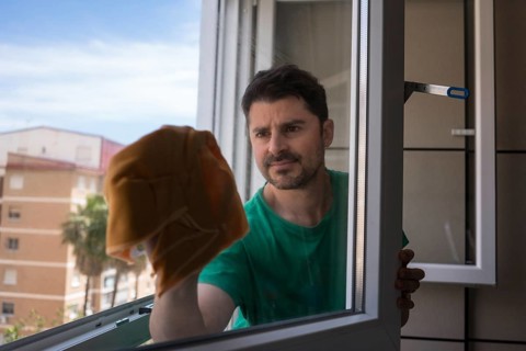 A man in casual attire carefully cleans a window in his urban apartment, ensuring clarity and