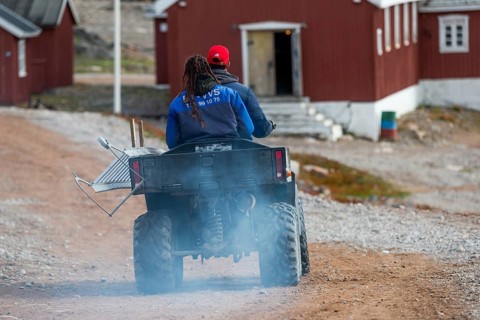 Two people on a quad bike or ATV driving on a dusty gravel road, remote Arctic Inuit settlement