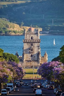 Blooming jacaranda trees line the street as Belem Tower rises above the Tagus River, with sailboats