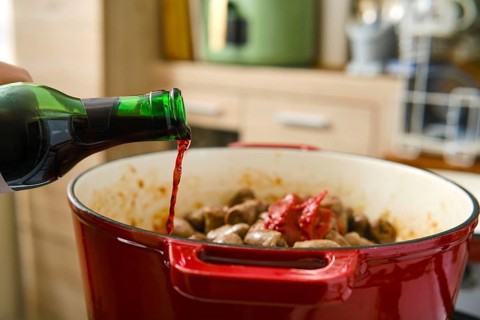 A person pours red wine into a pot filled with meat and sauce on a stove in a kitchen. The scene