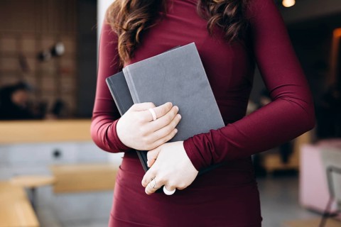 Female student in a maroon dress holding two gray books, standing in a contemporary study room with