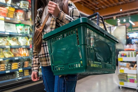 Woman carrying a green shopping basket, walking past refrigeration units in a supermarket, focusing