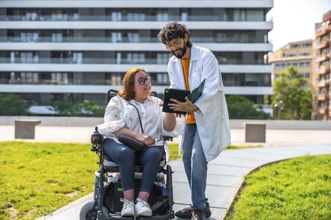 Doctor sharing information on a tablet with a smiling woman in a wheelchair, enjoying a sunny day