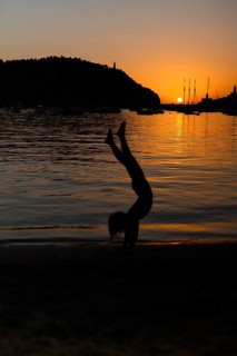 A child silhouette performs a handstand on a sandy beach as the sun sets over a tranquil sea,