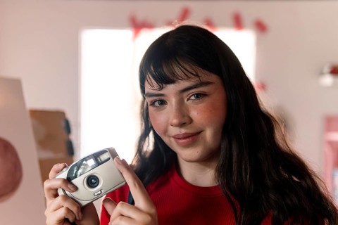 A young woman with long dark hair and a warm smile holds a vintage film camera. She wears a red