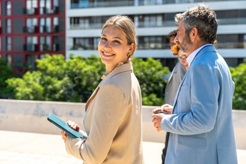 Businesswoman smiling and holding a tablet while walking outdoors with diverse colleagues in suits,