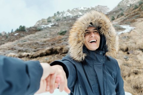A cheerful woman in a fur-lined hooded coat smiles while holding a hand, set against the snowy