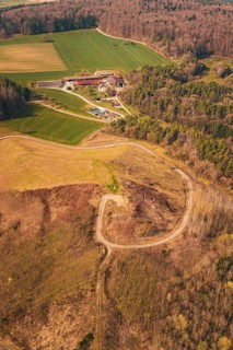 Aerial view of a hilly area with fields, a building and a winding path surrounded by forest, old