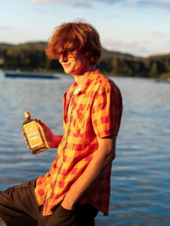 Young man standing by the lake, holding a bottle of alcohol in his hands, herbal liqueur, red hair,