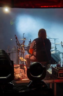 View from behind of a musician playing drums on stage, surrounded by atmospheric lights and fog,