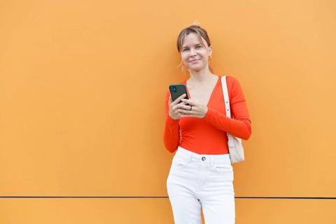 A woman stands casually against a bright orange wall in Warsaw, using a smartphone Dressed in a