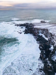 Aerial view of Iceland's rugged winter coastline meeting the sea. Snow-dusted cliffs create a