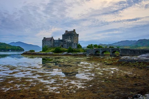 An old castle reflected in calm water, connected by a stone bridge under an evening sky, Eilean