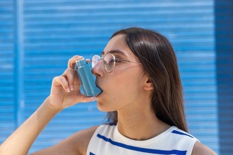A young woman uses an inhaler for asthma relief while standing against a bright blue background in
