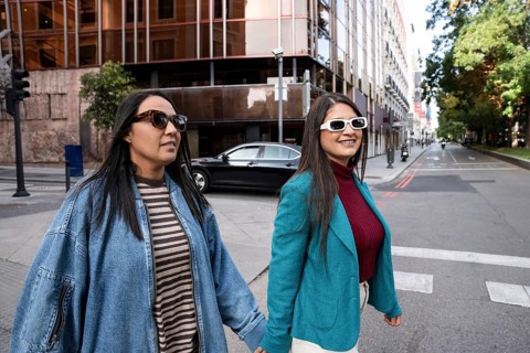 A joyful Latin lesbian couple walks hand in hand through a city street. They wear sunglasses and