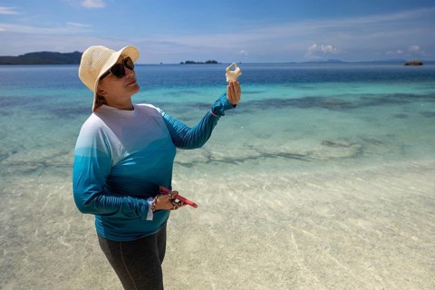 A woman wearing a sun hat and sunglasses holds a seashell while standing on a tropical beach of