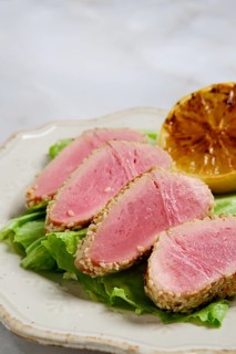 Sliced seared tuna rests on bed of lettuce with sesame crust. Grilled lemon half sits beside the