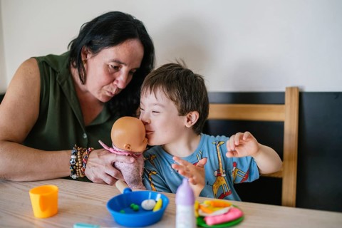 A young boy with Down syndrome plays joyfully with a doll, accompanied by an attentive woman. The