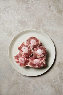 Five raw oxtail pieces are neatly arranged on a round, white ceramic plate. The marbled texture and
