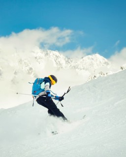 Cool skier performing switch skiing trick backwards on snowy slope with mountains in background