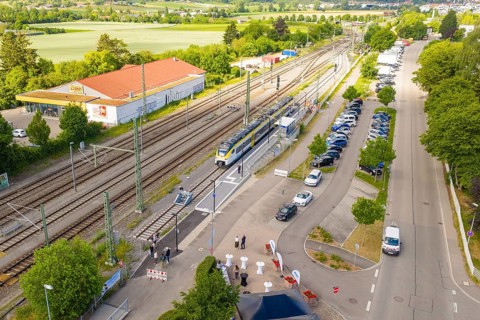 A train station with a stopping train, parked cars and people on a sunny street, inauguration of