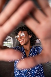 A vibrant woman with curly hair and glasses smiles, framed by her hands in a playful gesture,