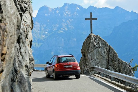 Red car passes a memorial cross on a bend in the narrow mountain road Route de Van in the Vallon de