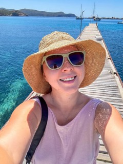 A young Caucasian woman with blonde hair smiles while standing on a wooden pier. She wears a straw