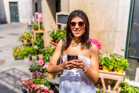 Young woman browsing her smartphone while enjoying a sunny day at a vibrant flower shop, surrounded