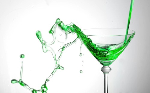 A powerful flood of green liquid pours into a cocktail glass and spills over, studio shot against a