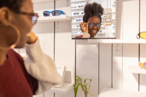 A woman enjoys selecting sunglasses in a trendy store. She admires her choice in the wall mirror,