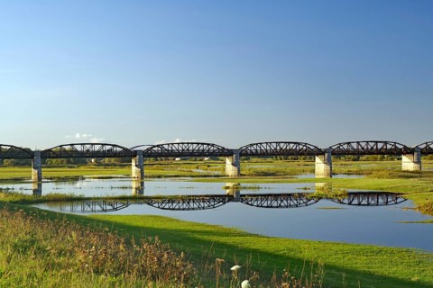 A bridge stretches over a calm body of water, surrounded by green meadows under a blue sky,