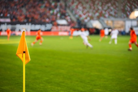 A close-up of a bright yellow corner flag on a grass pitch with a football match and stadium crowd