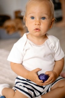 A toddler with bright blue eyes looks curiously at something, holding a small purple toy