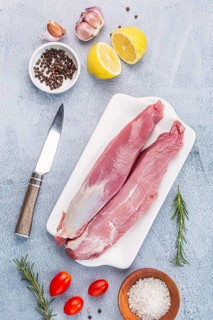 A fresh pork tenderloin on a white tray, surrounded by garlic, lemon, rosemary, tomatoes, salt,