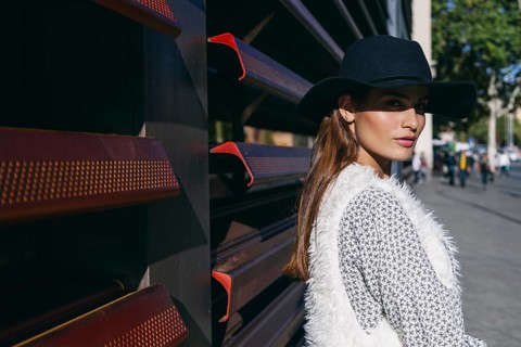 Woman wearing a stylish black hat and white vest, standing against a modern urban backdrop. The sun