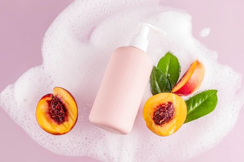 A pink shower gel bottle lies amidst soap bubbles, fresh peach halves, and glossy green leaves on a