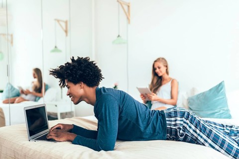 A young man using a laptop and a woman browsing a tablet in a cozy bedroom. They are casually