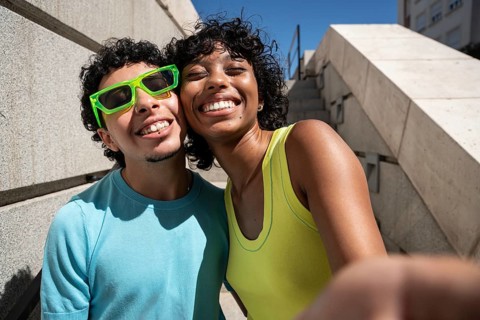 A joyful, multiethnic LGBTQ+ couple embraces and smiles warmly outdoors. Dressed casually, they