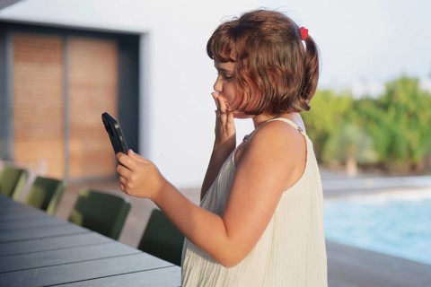 A young girl in a white dress enjoys a sunny summer day by the pool, engaging with her smartphone.
