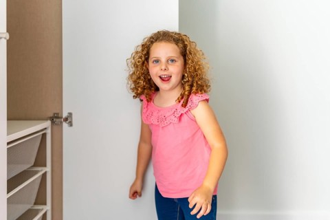 Little girl with curly red hair smiling cheerfully while exploring an empty closet with mesh