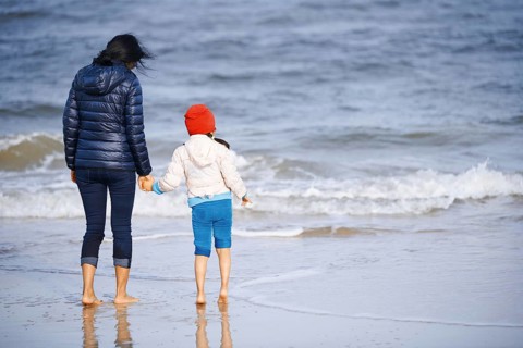 A mother and child, wearing warm clothing, hold hands while walking along a sandy beach in