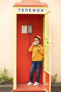 A preteen girl in a yellow jacket using a retro telephone inside a bright red phone booth with