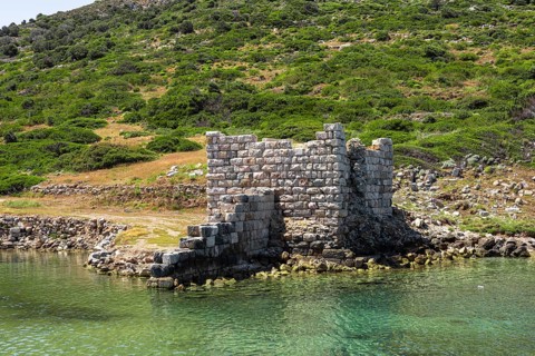 Ruins of an ancient stone structure by the water in Turkiye. Lush green hills surround the site,