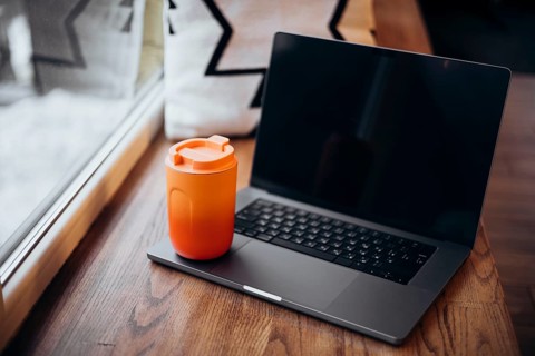 A modern laptop paired with a vibrant orange thermos cup sits beside a sunny window, illustrating a