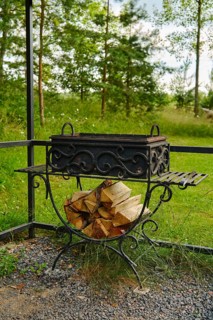 Fire pit stands in garden with neatly stacked firewood beside it. Tall trees and green grass